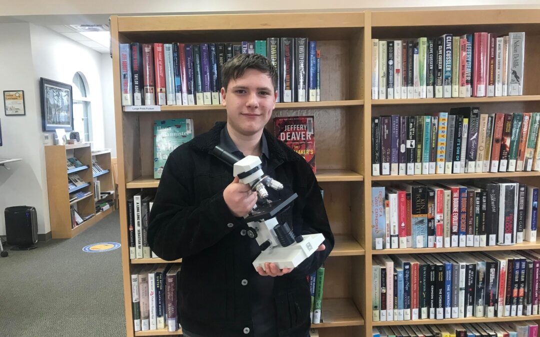 Photo of Sam Robinson holding a microscope in front to shelves of library books